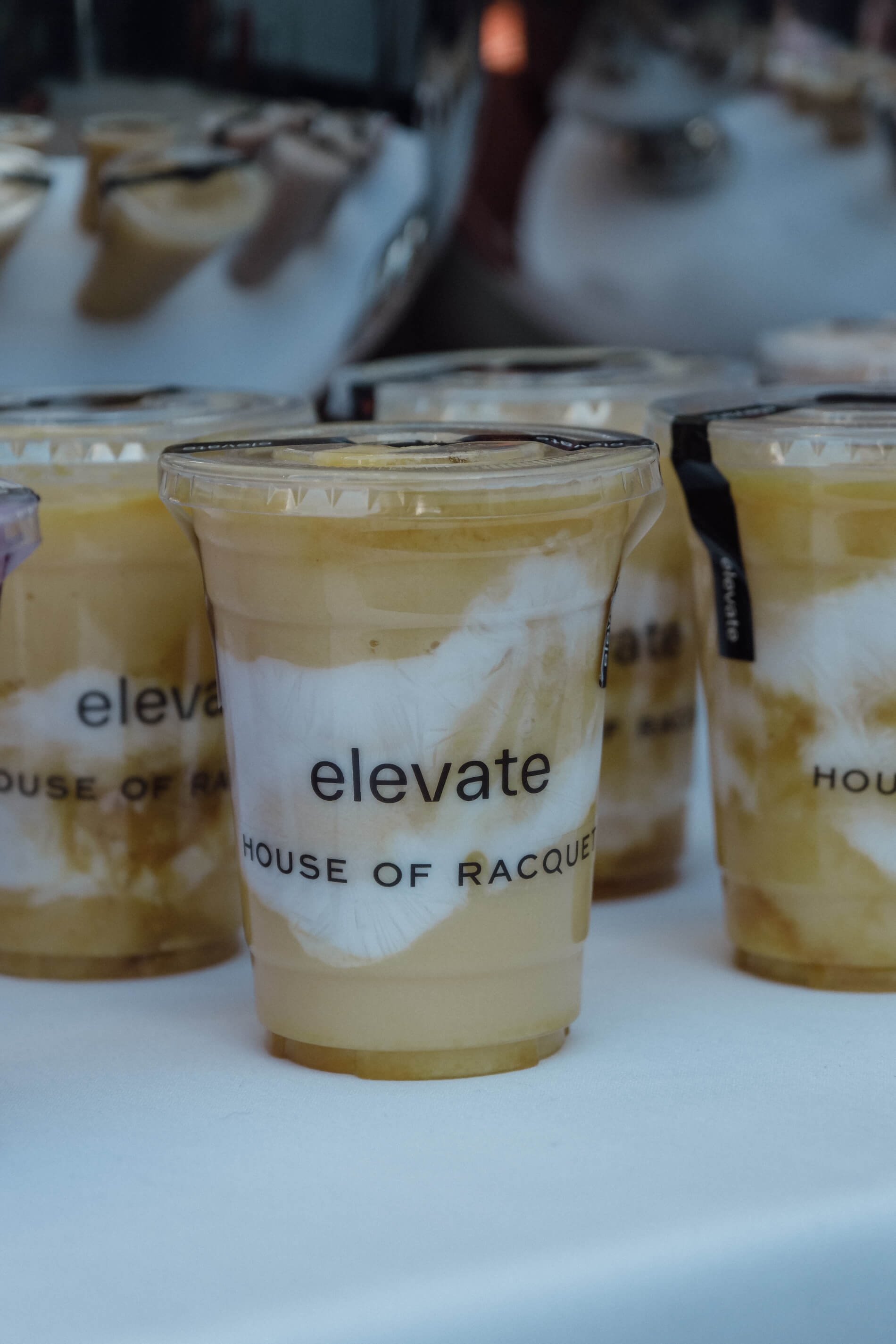 A group of iced coffee drinks in transparent plastic cups are aligned. The word 'Elevate' is printed in black letters at the front of each cup with 'House of Racquet' printed underneath.