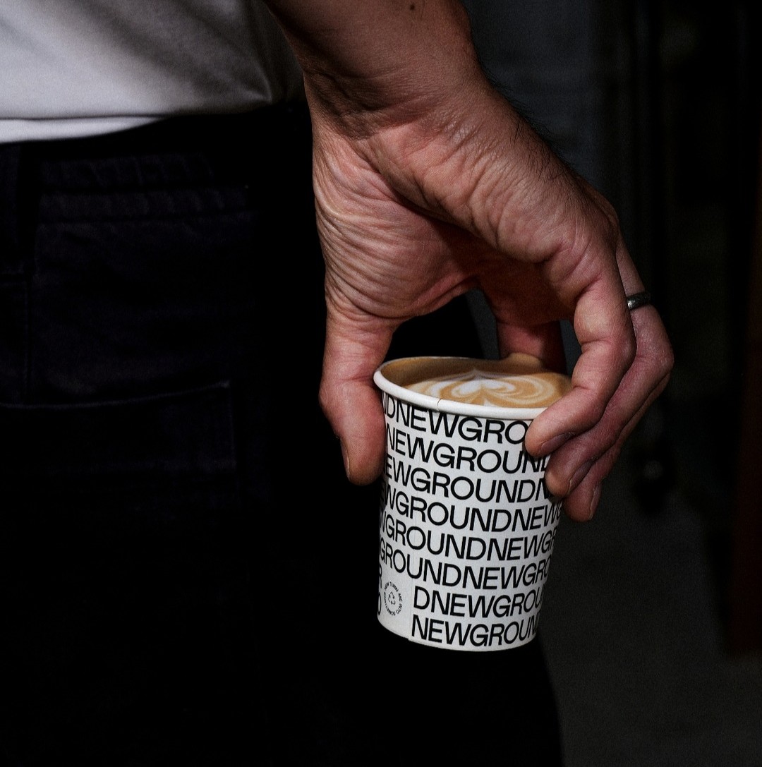 Close up of a hand holding a white Newground coffee paper cup with a latte inside.